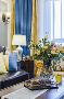 客厅色调柔和通透，以白色为主，搭配清新的蓝黄色打造了温馨的底色，配上柔软的黑沙发，恬雅沉稳而宁静。灿烂绽放的绿植，增添了空间的优雅情调与活力；